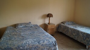 A room with two beds, each with a patterned bedspread, a wooden nightstand with a lamp, and a plain wall. - CABAÑA EN SIERRA DE LOBOS (León)
