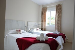 A hotel room with two beds, each with a red blanket and rolled towels. There is a window with curtains, a lamp, and a sign on the wall. - Apartment 50m from the sandy beach with communal pool in Denia, km 3 (Dénia)