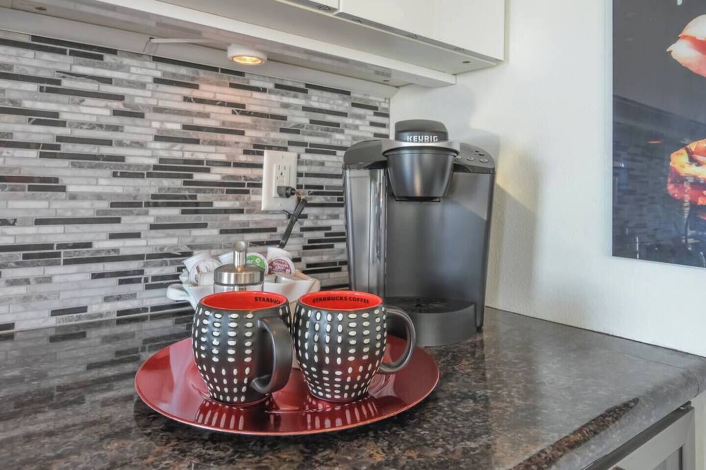 A kitchen with a modern design, featuring a coffee maker and two red mugs with white polka dots on a granite countertop.