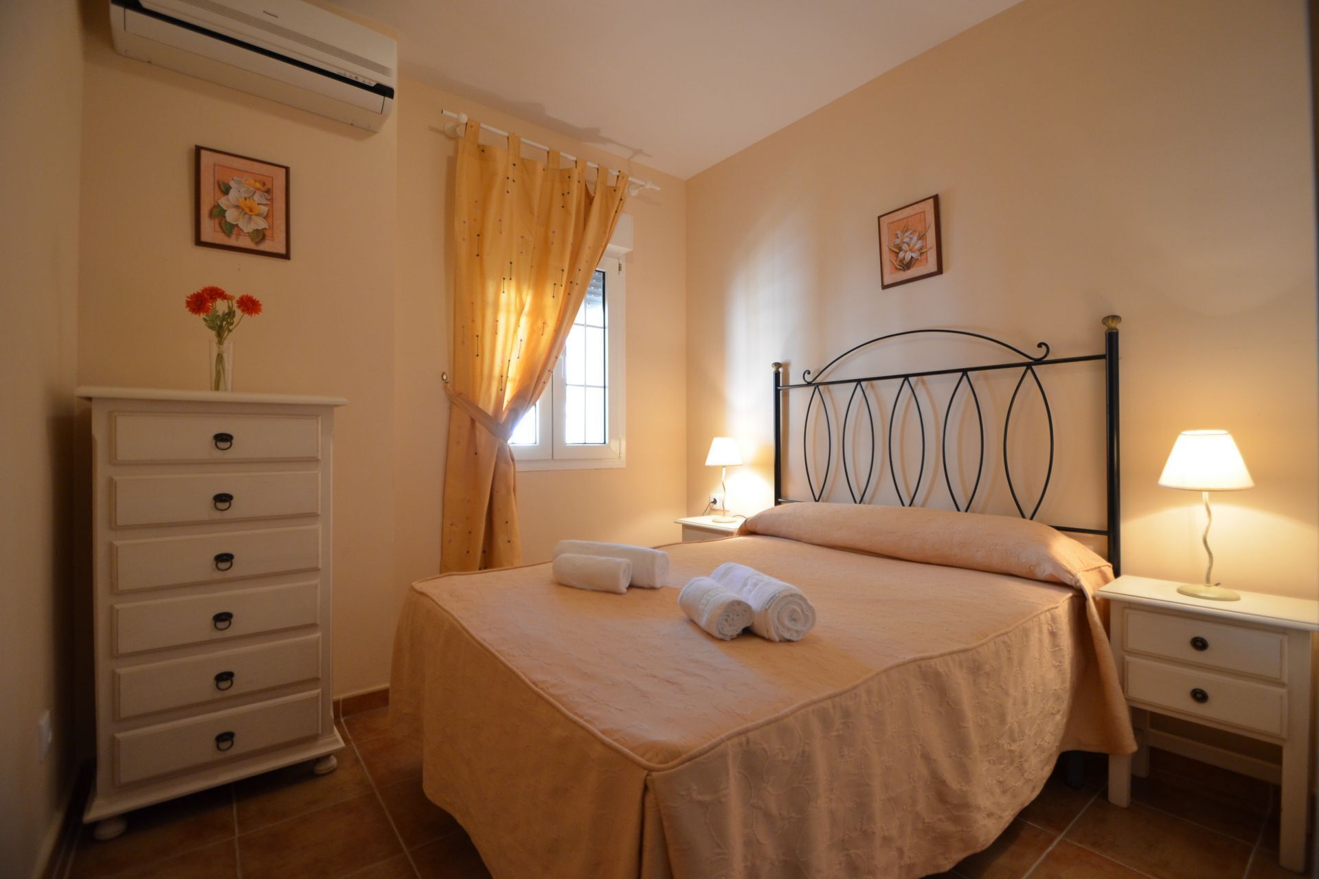 A bedroom with a bed, a dresser, two bedside tables with lamps, a window with curtains, and an air conditioning unit.