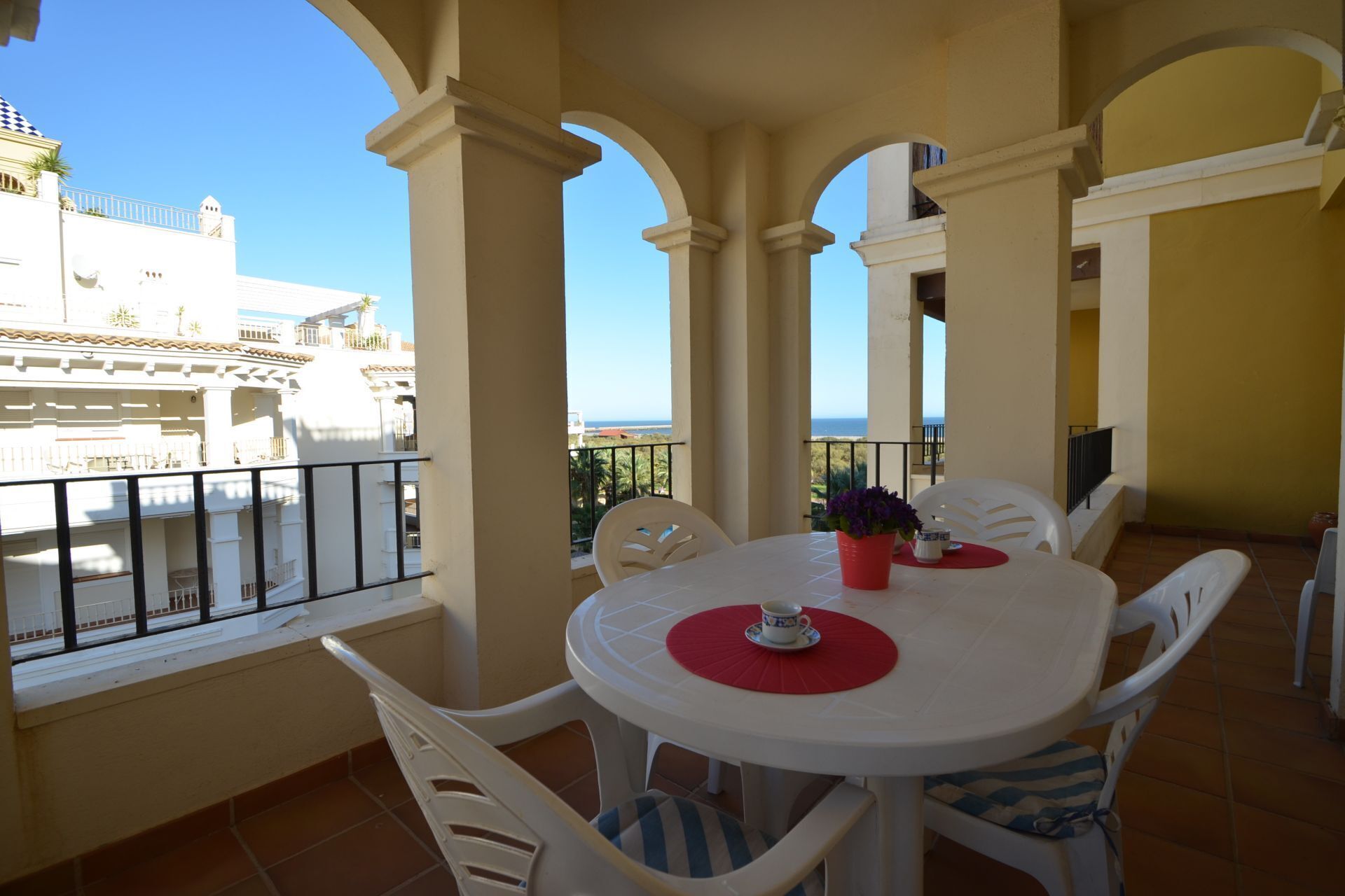 Un balcon avec une table et des chaises blanches, un set de table rouge et une plante en pot.