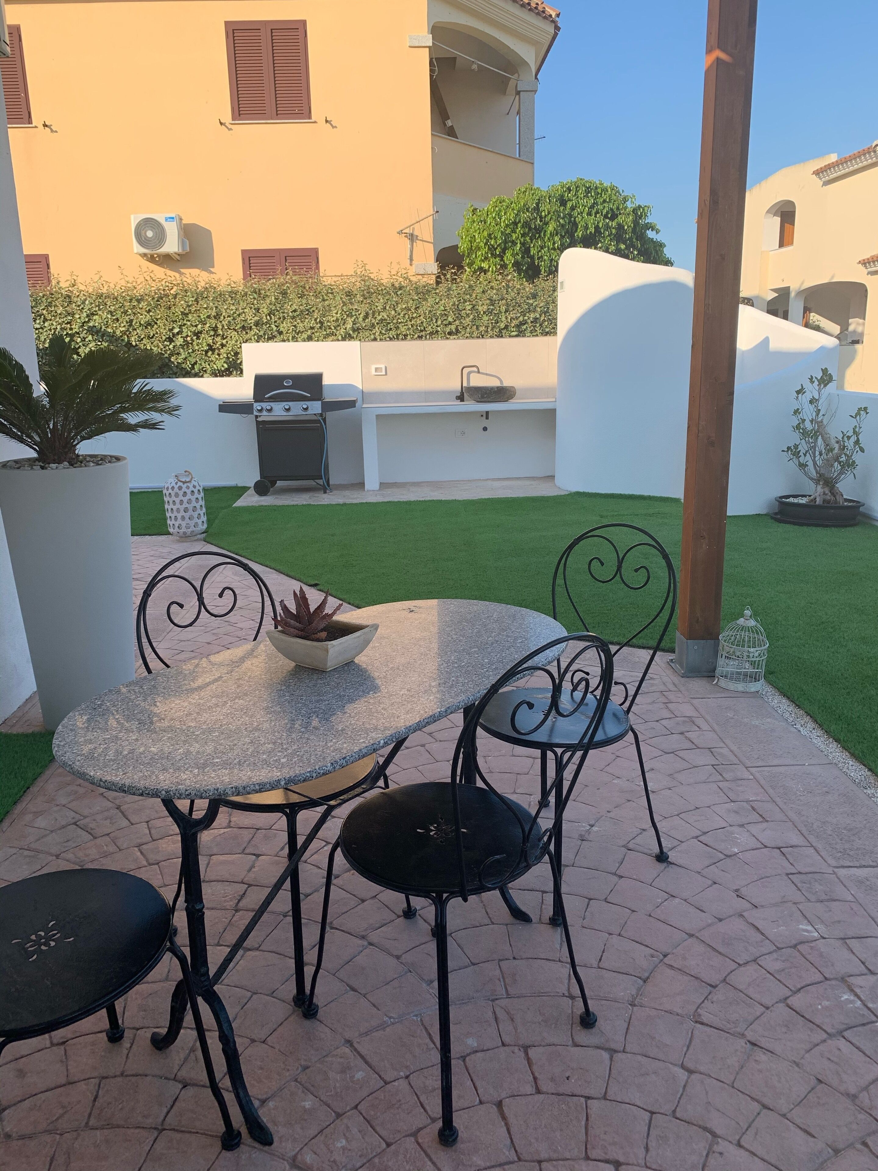 An outdoor patio with a round table and two chairs, a grill, and a small garden area with a potted plant.
