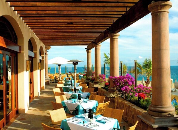 An outdoor dining area with tables and chairs, columns, and a view of the ocean.