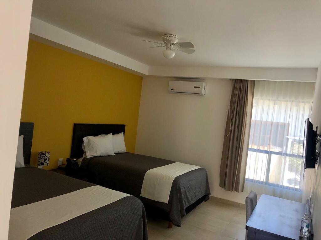 Habitación de hotel con dos camas, un ventilador de techo, un aire acondicionado, una ventana con cortinas y una pequeña mesa con una silla.