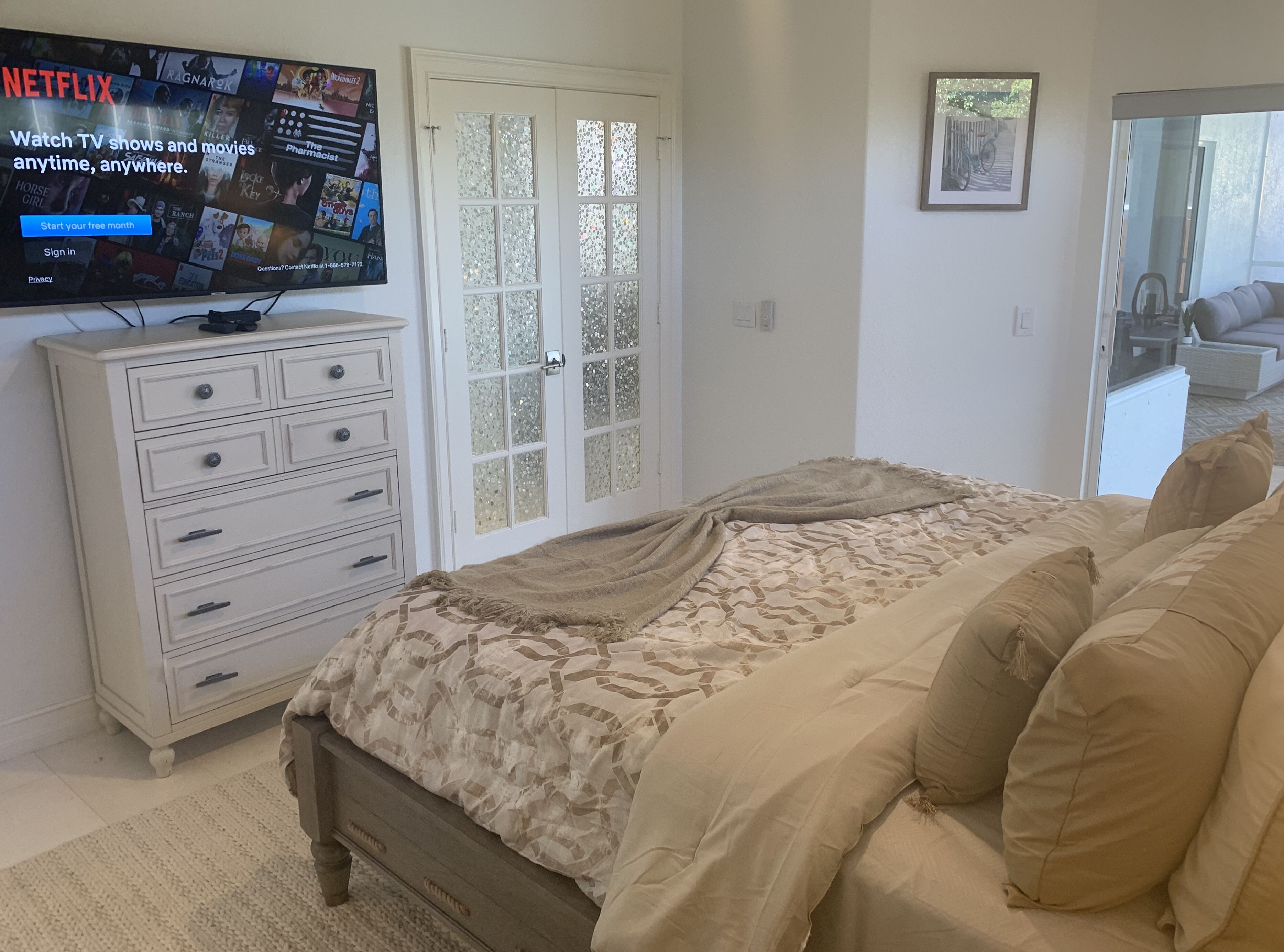 Une chambre à coucher comprenant un lit, une commode, un téléviseur affichant l’interface de Netflix et une porte dotée de panneaux en verre dépoli.