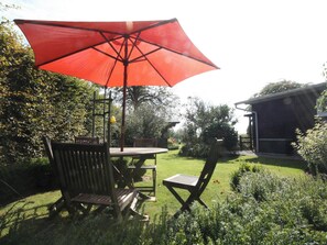 A patio with a wooden table and chairs, a red umbrella, and a garden with various plants and a shed in the background. - Cozy Chalet With Pond, Malmedy (Malmedy)