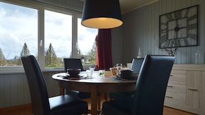 Un comedor con una mesa redonda, cuatro sillas, una lámpara colgante, un reloj en la pared y vistas a árboles y una casa por la ventana.