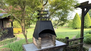 Un horno de pizza de piedra con techo negro, rodeado por una pérgola de madera y abundante vegetación.