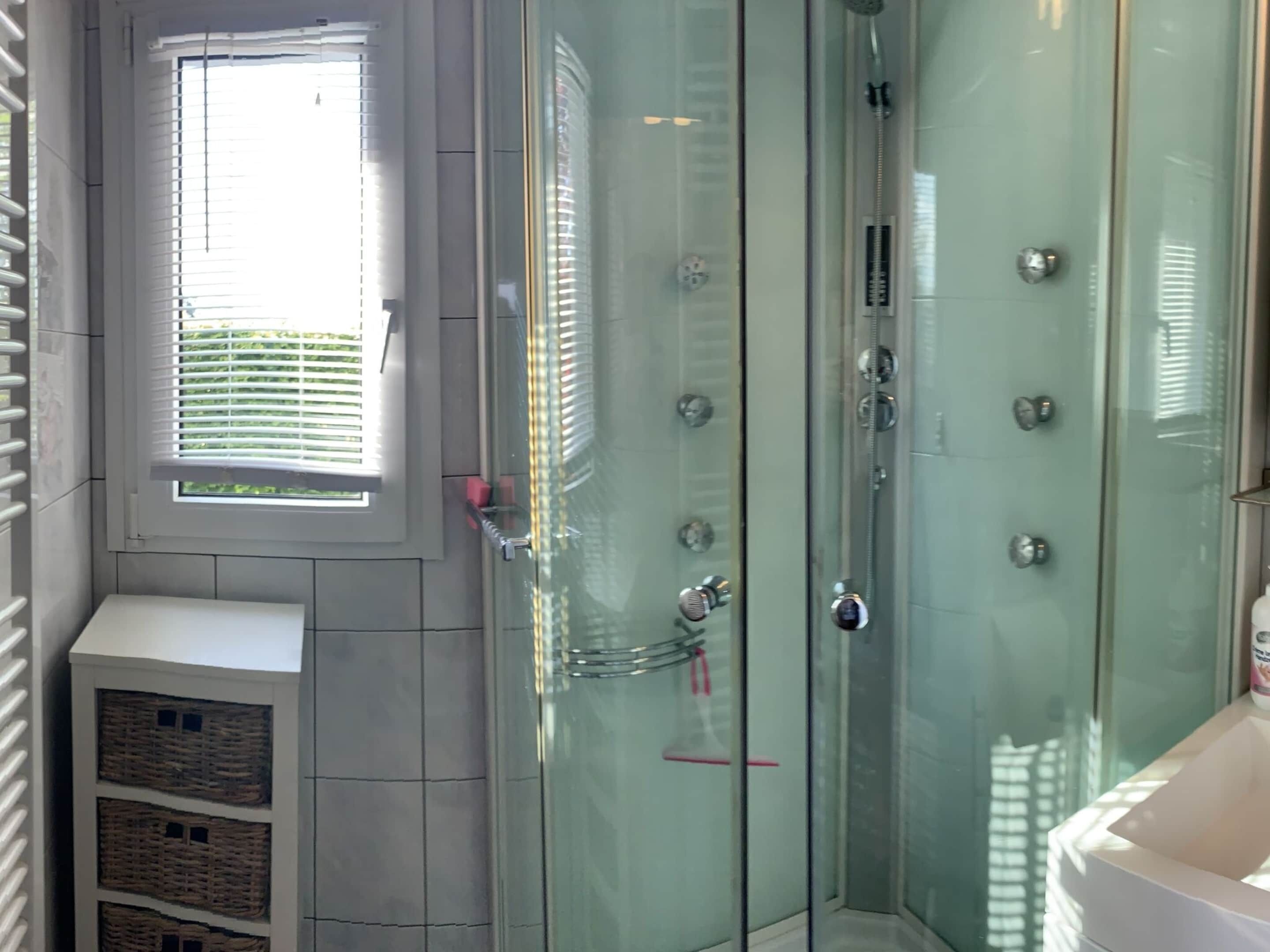 A glass-enclosed shower with multiple controls, a window with blinds, and a white cabinet with baskets.