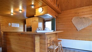 Interior de uma cabana de madeira com cozinha, uma decoração em formato de coração na parede e um banquinho de madeira.