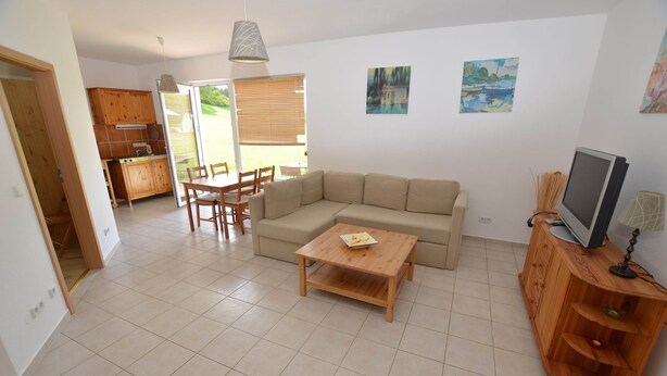 A spacious living area with a sofa, a coffee table, and a TV on a wooden cabinet. There is a dining area with chairs and a table, and a kitchen with wooden cabinets and a window. - Retreat With Garden Views (Dolní Lánov)