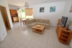 A spacious living area with a sofa, a coffee table, and a TV on a wooden cabinet. There is a dining area with chairs and a table, and a kitchen with wooden cabinets and a window. - Retreat With Garden Views (Dolní Lánov)