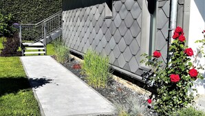 A building with a grey tiled exterior, a metal staircase leading to a door, and a garden with various plants and red flowers. - Apartment in Weywertz Near Ski Slopes (Buetgenbach)