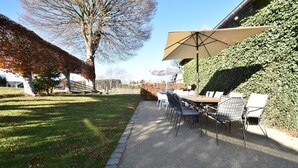 Un patio al aire libre con una mesa, sillas, un paraguas grande y un seto.