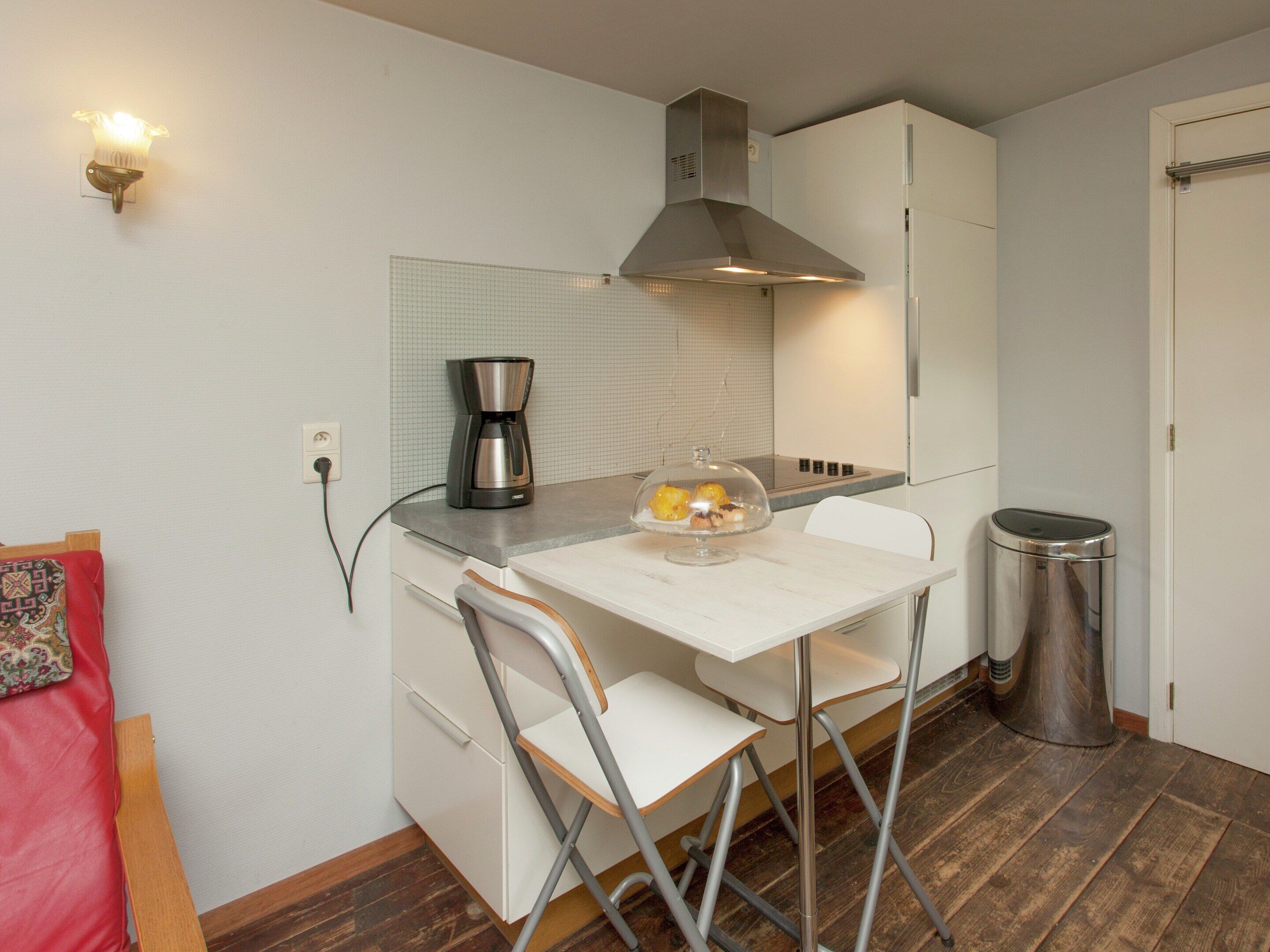A compact kitchen with a coffee maker, a glass bowl with food, a trash can, and a foldable chair.