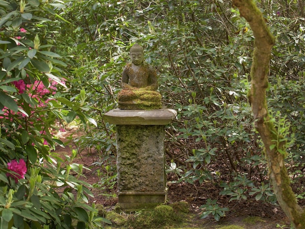 Una estatua de Buda de piedra en un jardín, rodeada de exuberante follaje y flores rosadas.