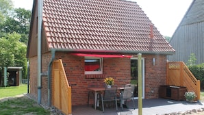 Een klein, wit vakantiehuis met pannendak, een terras met tuinmeubilair en een houten vlonder.