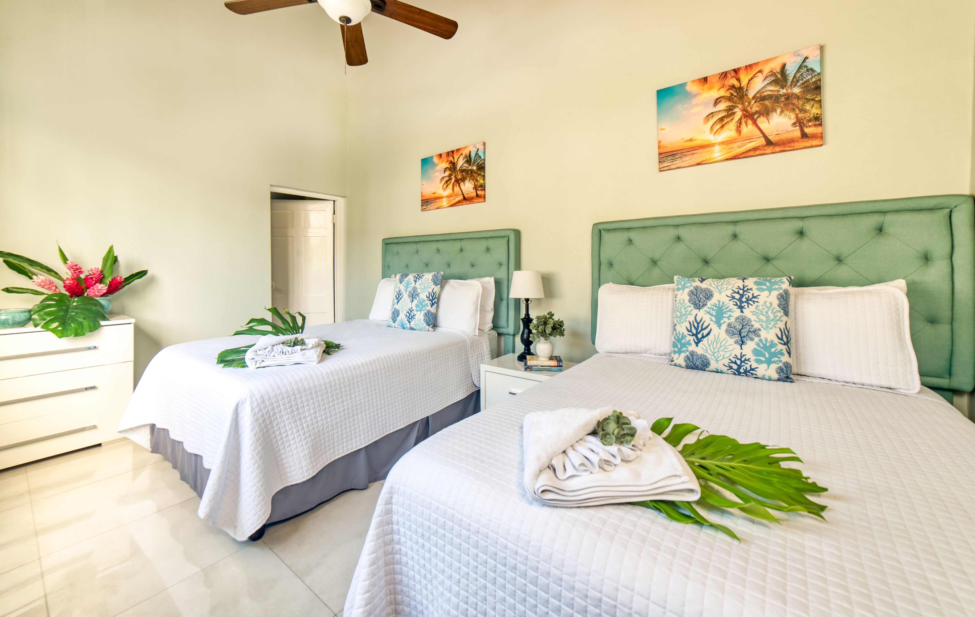 A hotel room with two beds, a ceiling fan, and artwork depicting tropical scenes.