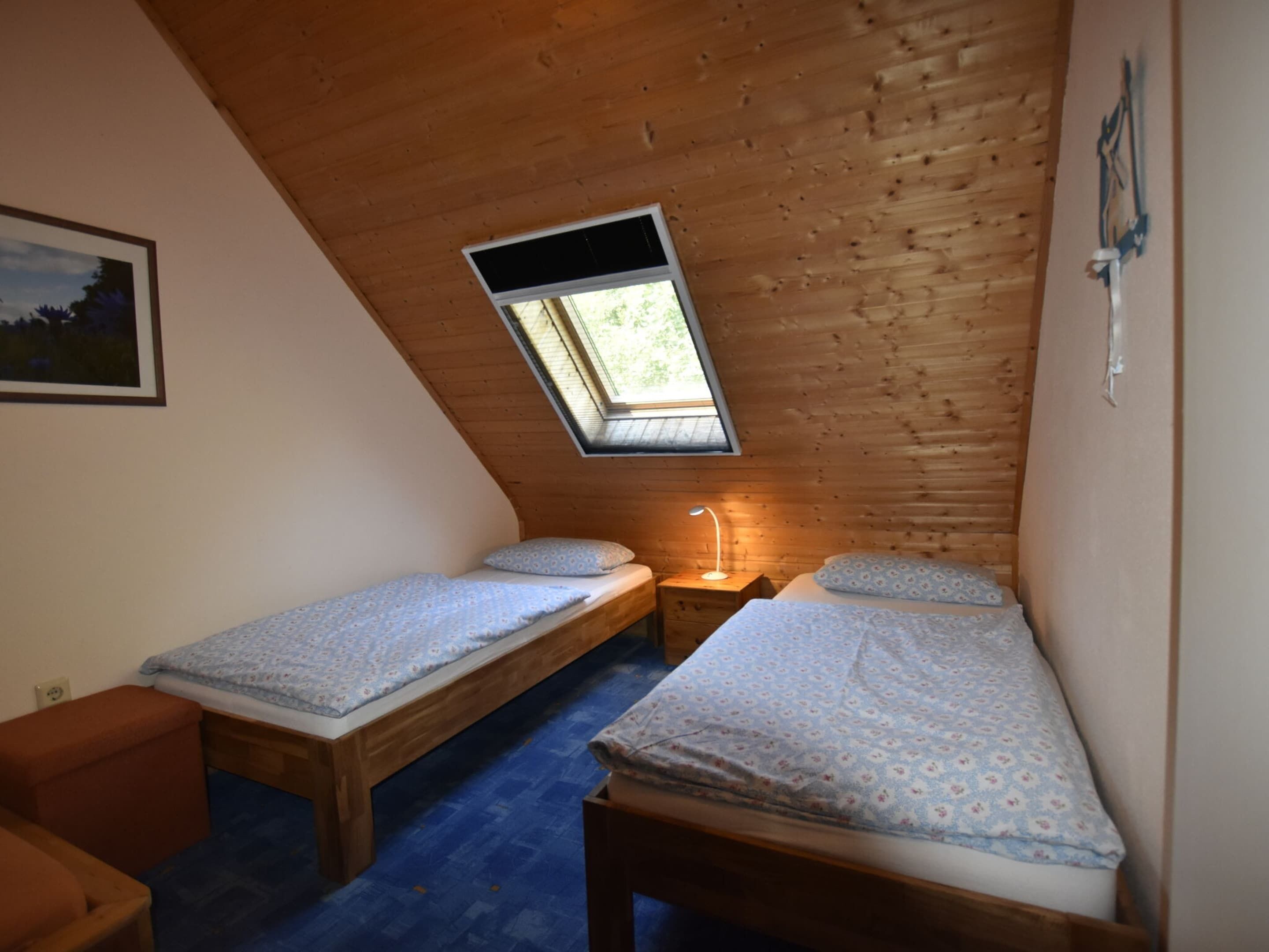 Una habitación pequeña y acogedora con dos camas de madera, un tragaluz y un escritorio con una lámpara.