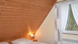 Et soveværelse med skrå træloft, en seng med hvide sengetøj, en sengetøjslampe og et vindue med lette gardiner.
