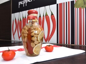 Um frasco de vidro cheio de nozes e fatias de limão sobre uma superfície branca, com tomate e uma parede com tema de pimentão ao fundo.