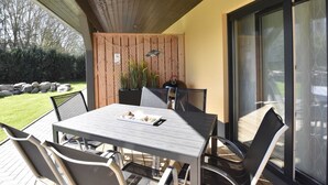 Un patio al aire libre con una mesa y sillas, techo de madera, puerta de cristal y vista al jardín.