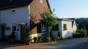 Une maison à deux étages avec un toit en pente, un petit garage attenant et une véranda à l’avant, agrémentée de plantes en pot.