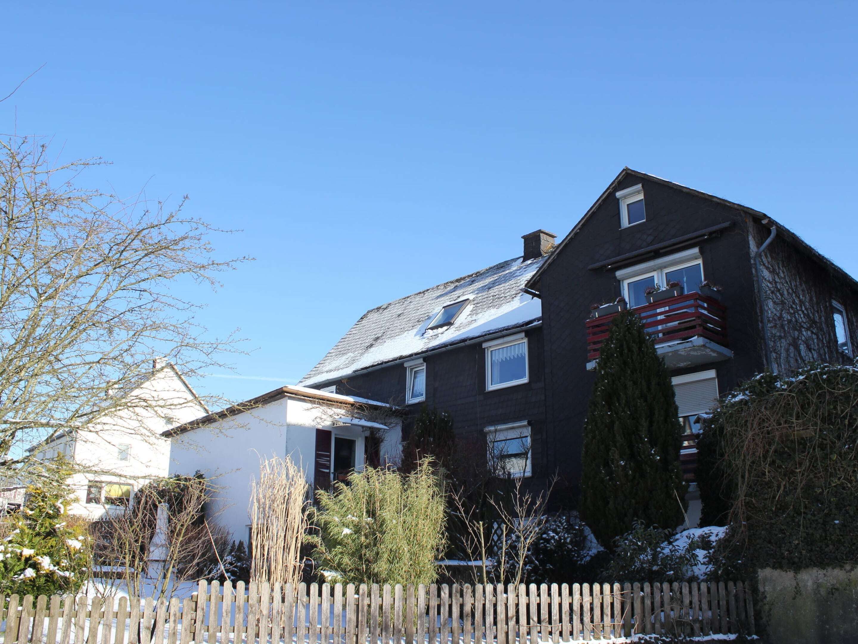 En tvåvånings bostadsbyggnad med en mörk fasad, en synlig balkong med röda räcken och en klarblå himmel.