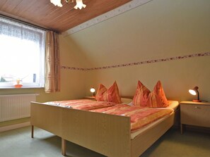 A bedroom with a wooden bed, two bedside lamps, a window with curtains, and a radiator. - Holiday Home With Terrace in the Harz Mountains (Harzgerode)