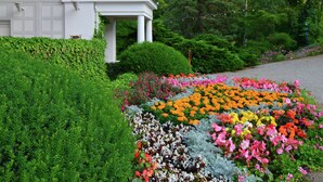 Ein weißes Gebäude mit einer Veranda, umgeben von üppiger Bepflanzung und einem farbenfrohen Blumenbeet.