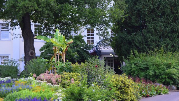 Ein Garten mit verschiedenen Blumen und Pflanzen, einem Kiesweg und einem Gebäude mit dem Schriftzug 'Baden'.