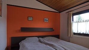 Et soveværelse med en orange væg, træloftsbrædder, en seng med stribet sengetøj og et vindue med gardiner.