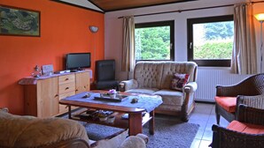 En hyggelig stue med træloft, et fjernsyn på et træskab, en komfortabel lænestol, en fletsofa og et stort vindue med gardiner.