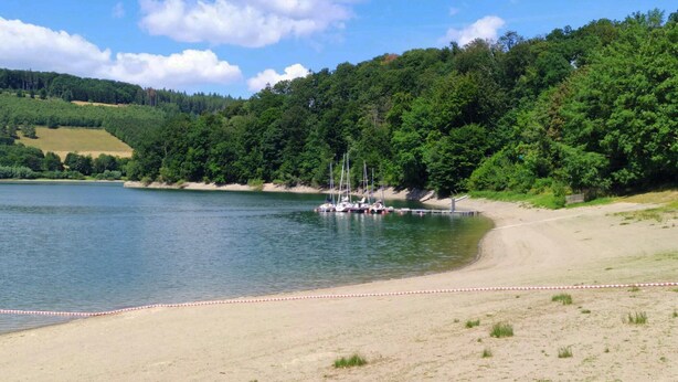Ein See mit Sandstrand und angestaubten Booten, umgeben von bewaldeten Hügeln.