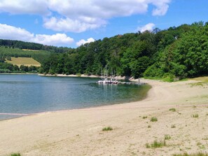 Une plage au bord d’un lac, avec des bateaux amarrés, entourée de collines boisées.
