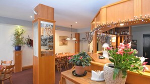 A cozy room with a wooden reception desk, a dining area with chairs, a potted plant, and a bookshelf.
