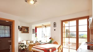 Uma área de refeições com uma mesa posta para dois, uma cadeira de madeira, um vaso com flores e vista para o exterior através de uma porta de vidro.