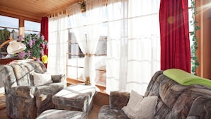 Quarto aconchegante com teto de madeira, janelas amplas com cortinas leves e vista para o exterior. Há dois sofás e uma mesinha com um vaso de flores.