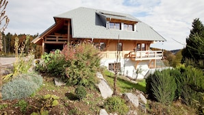 Uma casa de madeira de dois andares, com telhado inclinado, varanda e jardim com diversas plantas e pedras.