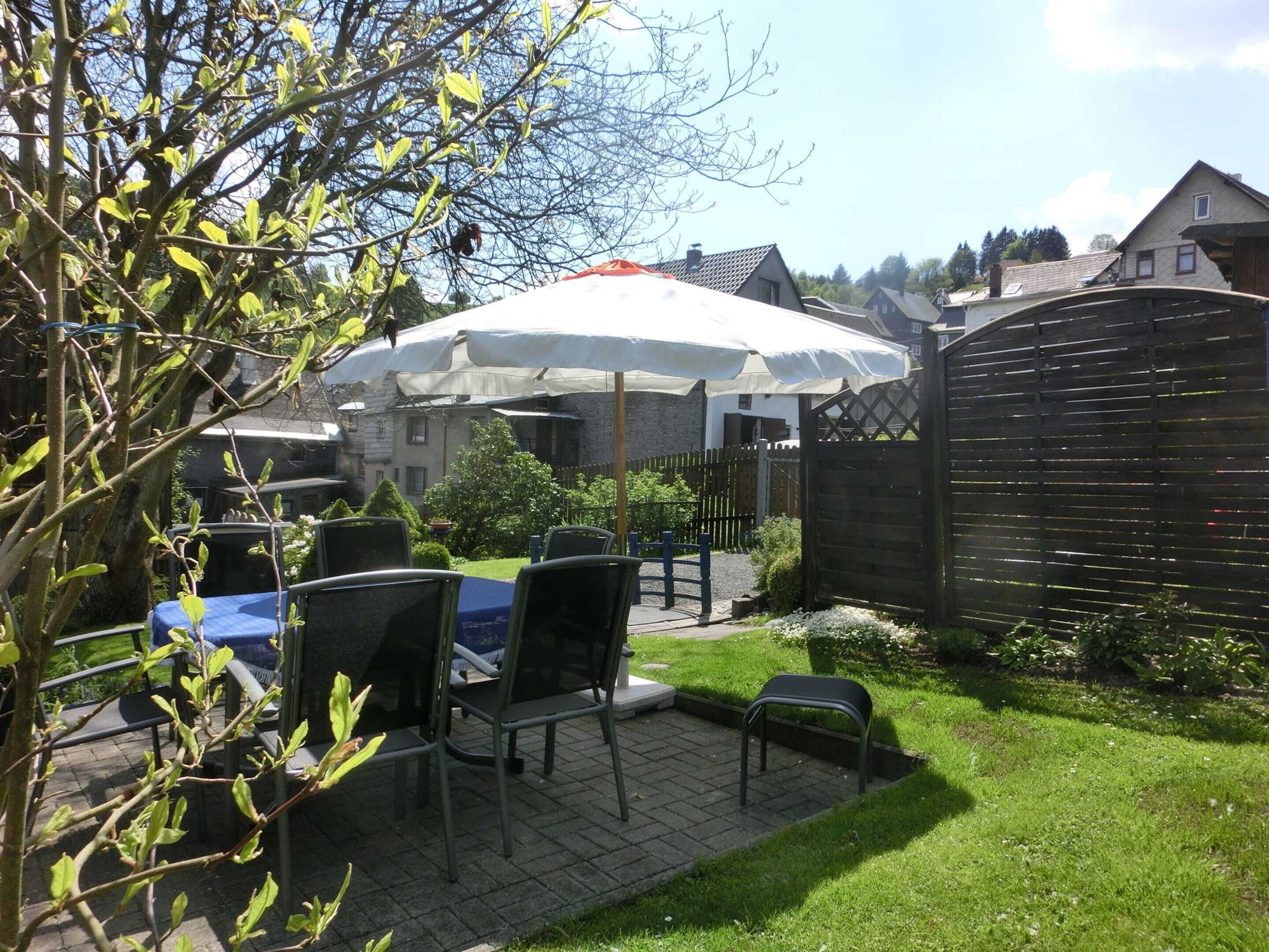 A patio area with a table and chairs under a white umbrella, surrounded by a wooden fence and greenery.