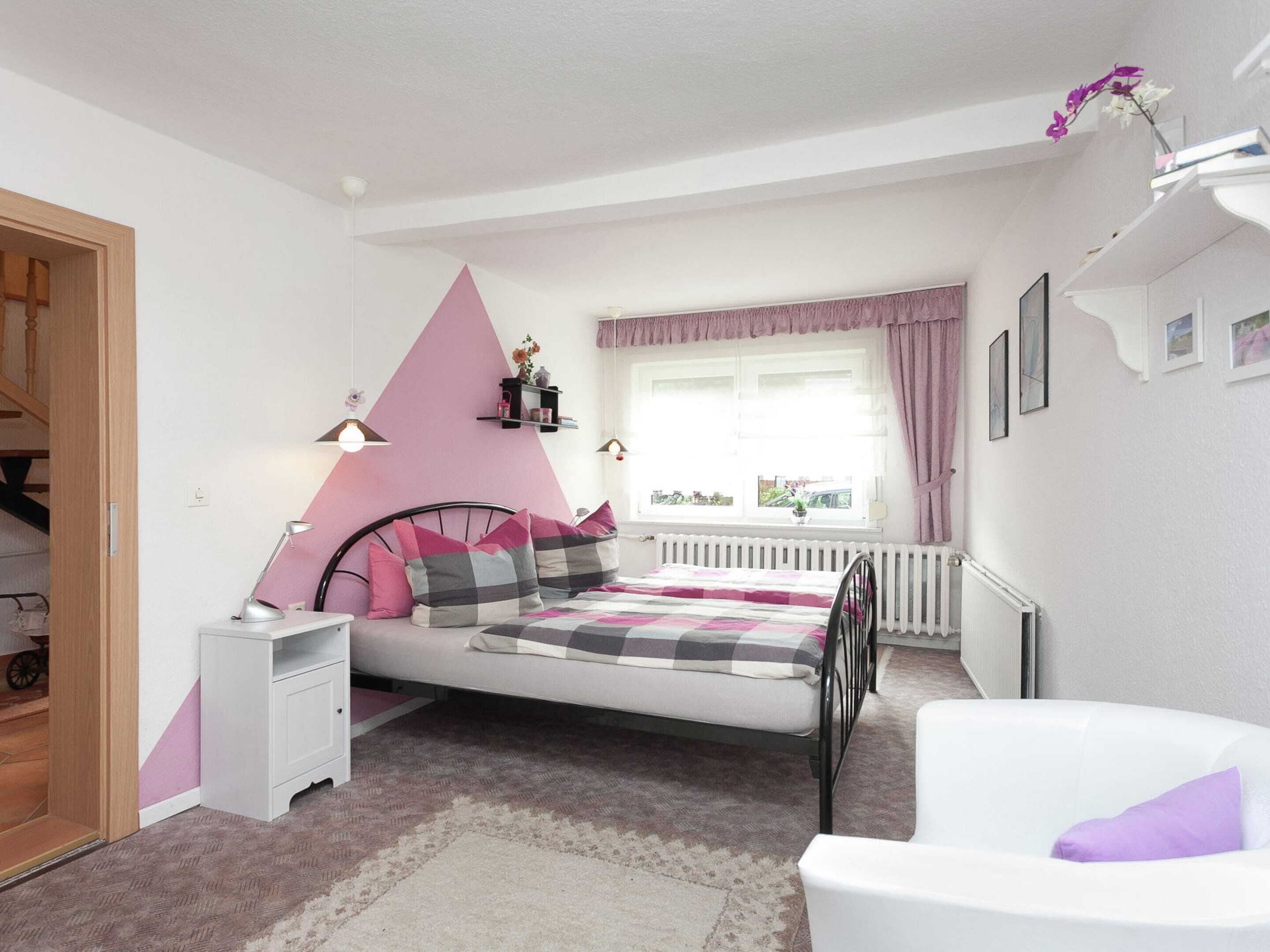 A bedroom with a bed, bedside table, chair, radiator, window, and a pink wall design.