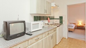 Una cocina compacta con televisión, microondas y cafetera. Se ve una cama y una mesita de noche con lámpara en la habitación contigua.