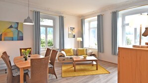 Una sala de estar moderna con una mesa de comedor de madera, sillas de mimbre, un sofá con cojines amarillos, una mesa de centro y ventanas grandes con cortinas.
