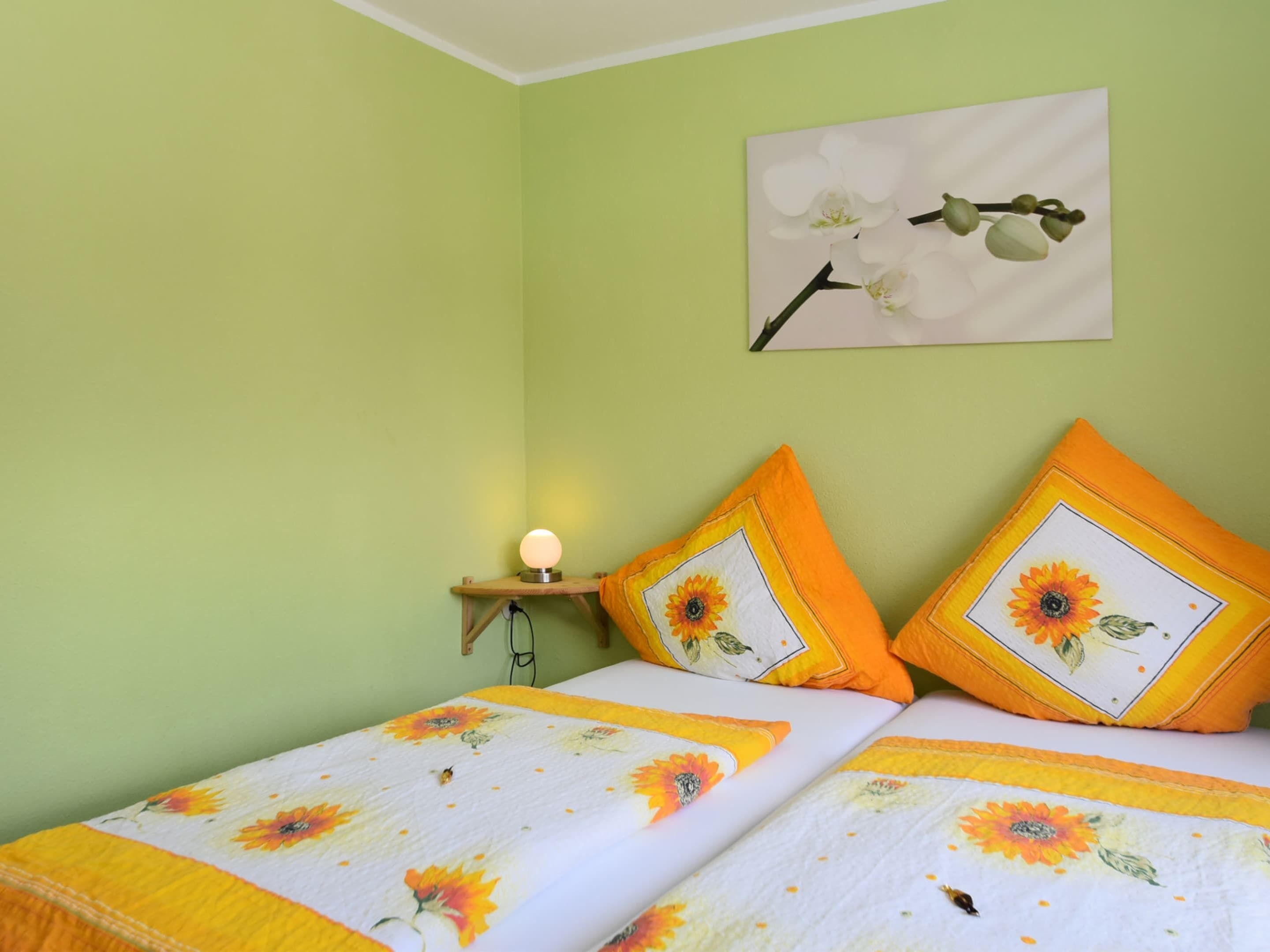 A bedroom with two beds, each with a floral duvet cover and matching pillows. There is a bedside table with a lamp, and a wall-mounted picture of flowers.