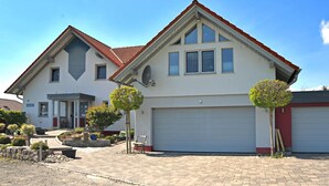 A two-story house with a red roof, a garage, and a driveway with trees and a bench. - Ferienhaus in Obernburg am Edersee (Voehl)