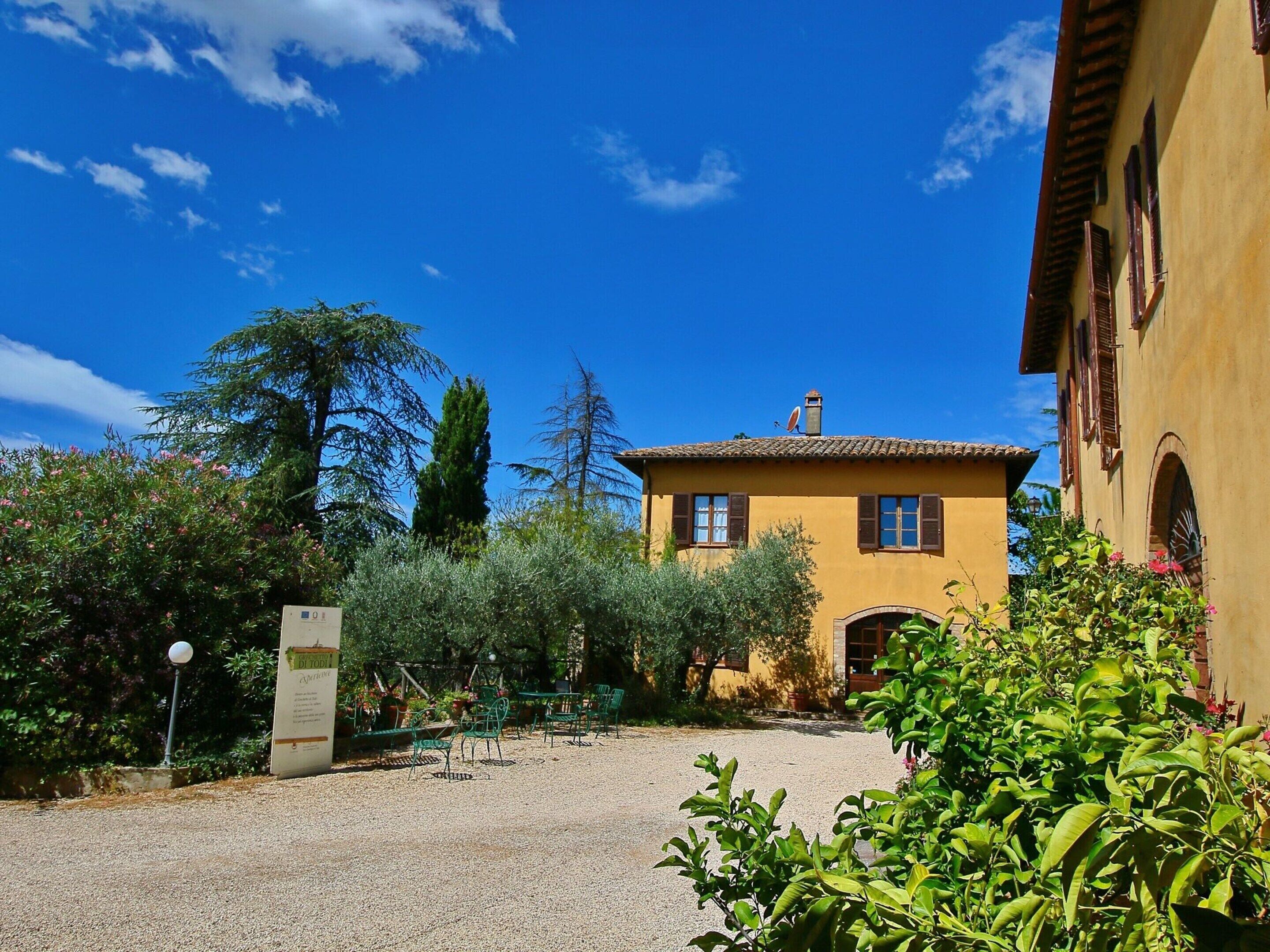 A yellow building with a tiled roof, surrounded by greenery and a gravel path.