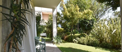A covered patio with a chair and table, surrounded by greenery and a lawn.