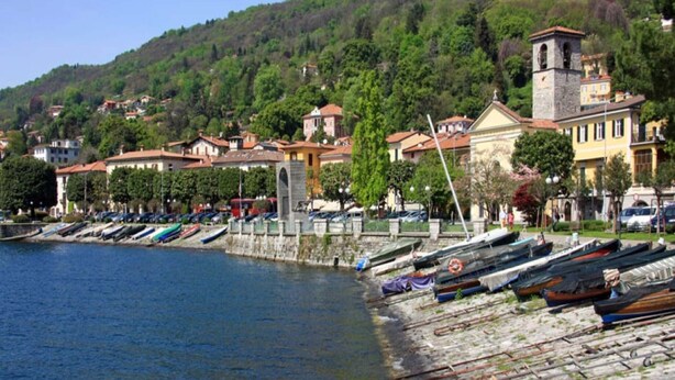 Une ville côtière où des bateaux sont amarrés le long de la rive, avec des bâtiments en pierre et une colline en arrière-plan.
