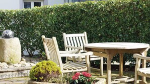 A wooden outdoor seating area with a round table and two chairs, surrounded by a stone planter with plants and a hedge in the background. - Holiday Home Fabryshof in Bollendorf (Bollendorf)