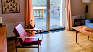 A room with a wooden floor, a red chair, a blue sofa, a table, a lamp, a picture frame, and a sliding glass door leading to an outdoor deck. - Stay With Garden in the Hunsruck (Stipshausen)